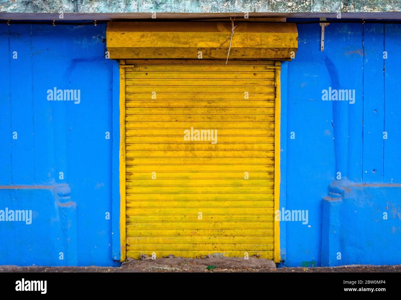 Natural street side frame of bright yellow closed shutter of a shop and bright blue walls on either side creating colour contrast. Stock Photo