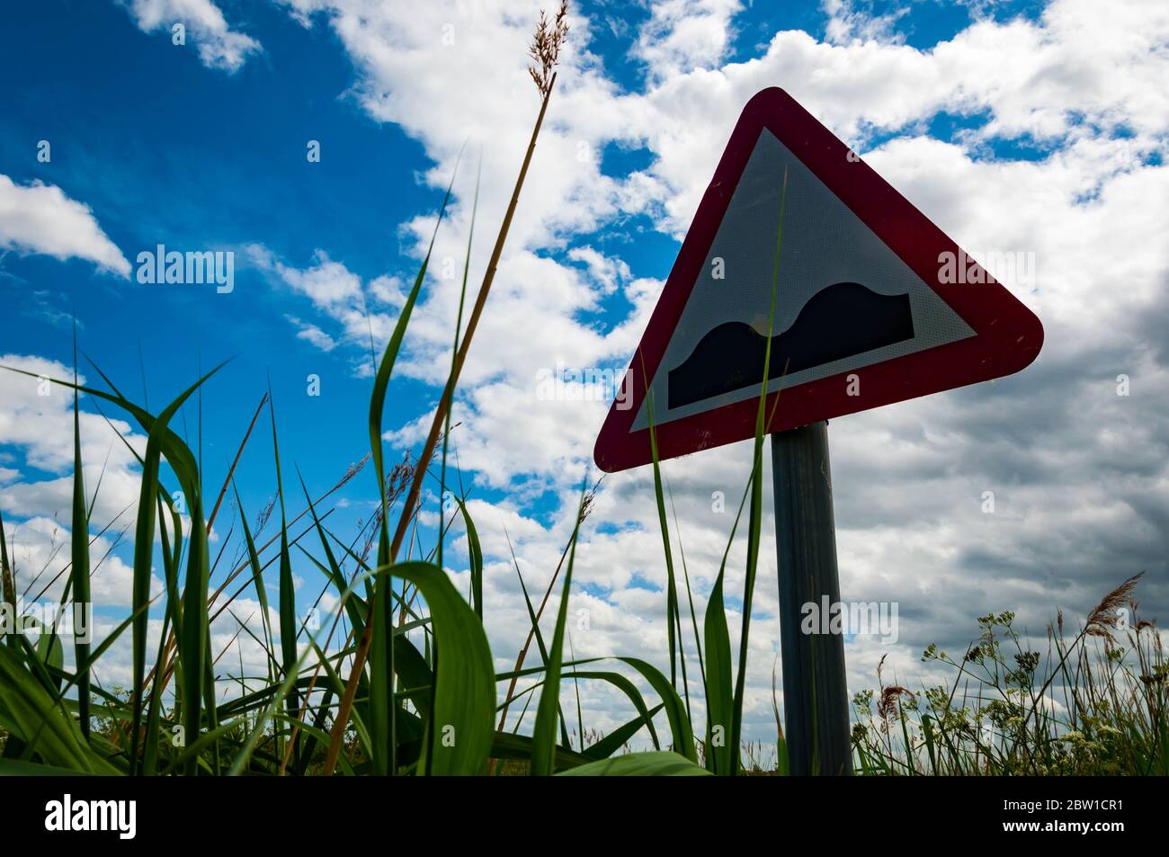 Bumpy road ahead sign in tall grass and bright clouds and blue sky in background Stock Photo