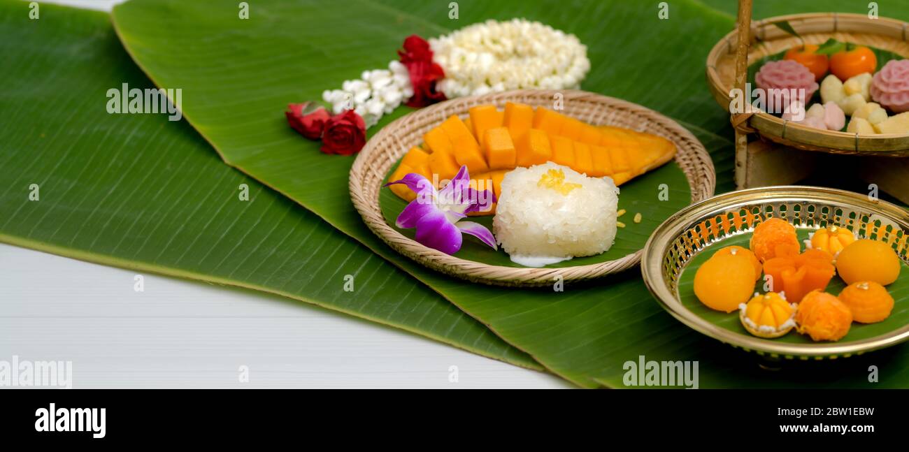 Close up view of several Thai traditional dessert serving on brass tray and wooden tray with jasmine garland and banana leaves decorated on white tabl Stock Photo
