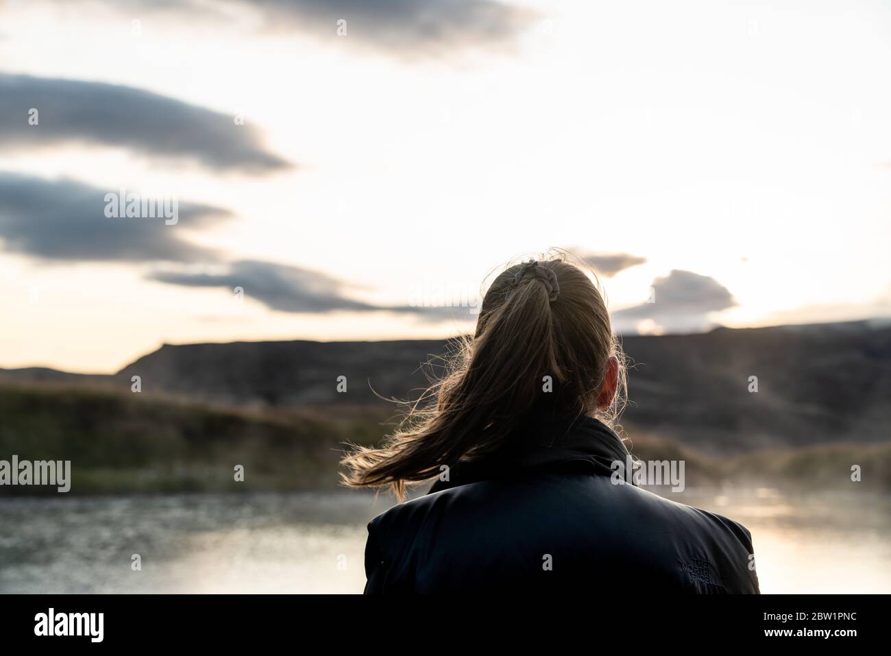 Young girl with brown hair in a ponytail gazing over a lake and a mountain into the sunset. Her hair blow in the wind and the sunlight contours the po Stock Photo
