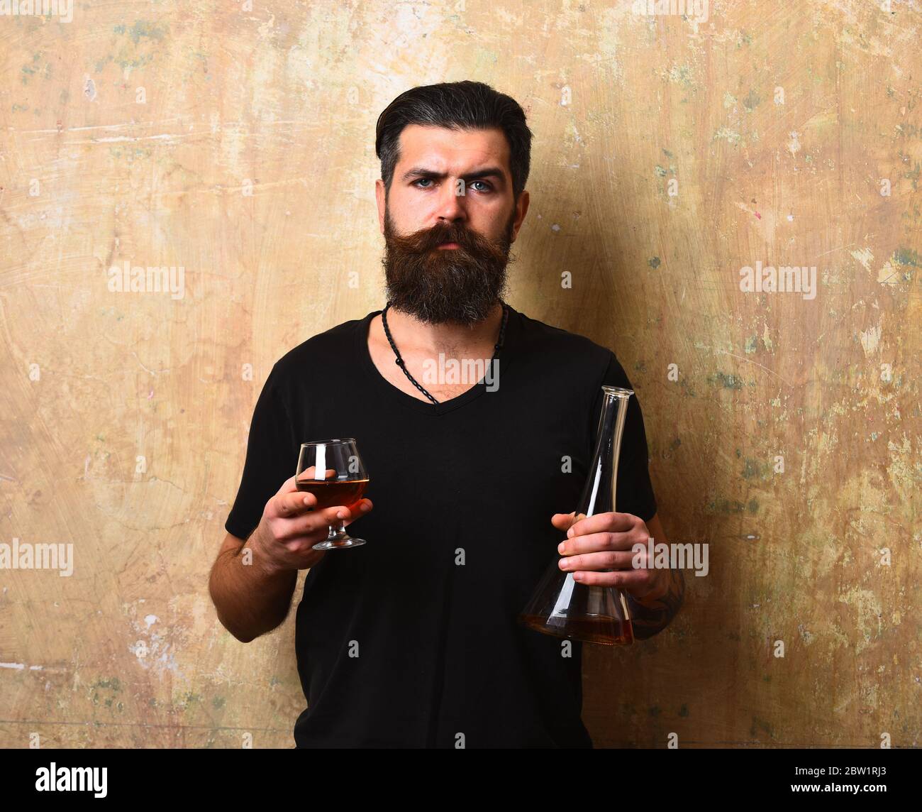 Man with beard and mustache holds alcoholic beverage on beige wall background. Service and catering concept. Guy with glass and bottle of cognac. Macho with serious face drinks brandy or whiskey. Stock Photo