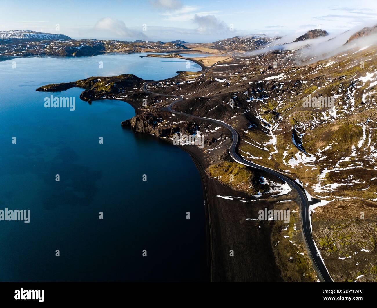 Aerial photograph of a dreamlike landscape with cloud covered mountains, a blue lake, and a small road meandering though the mountains Stock Photo