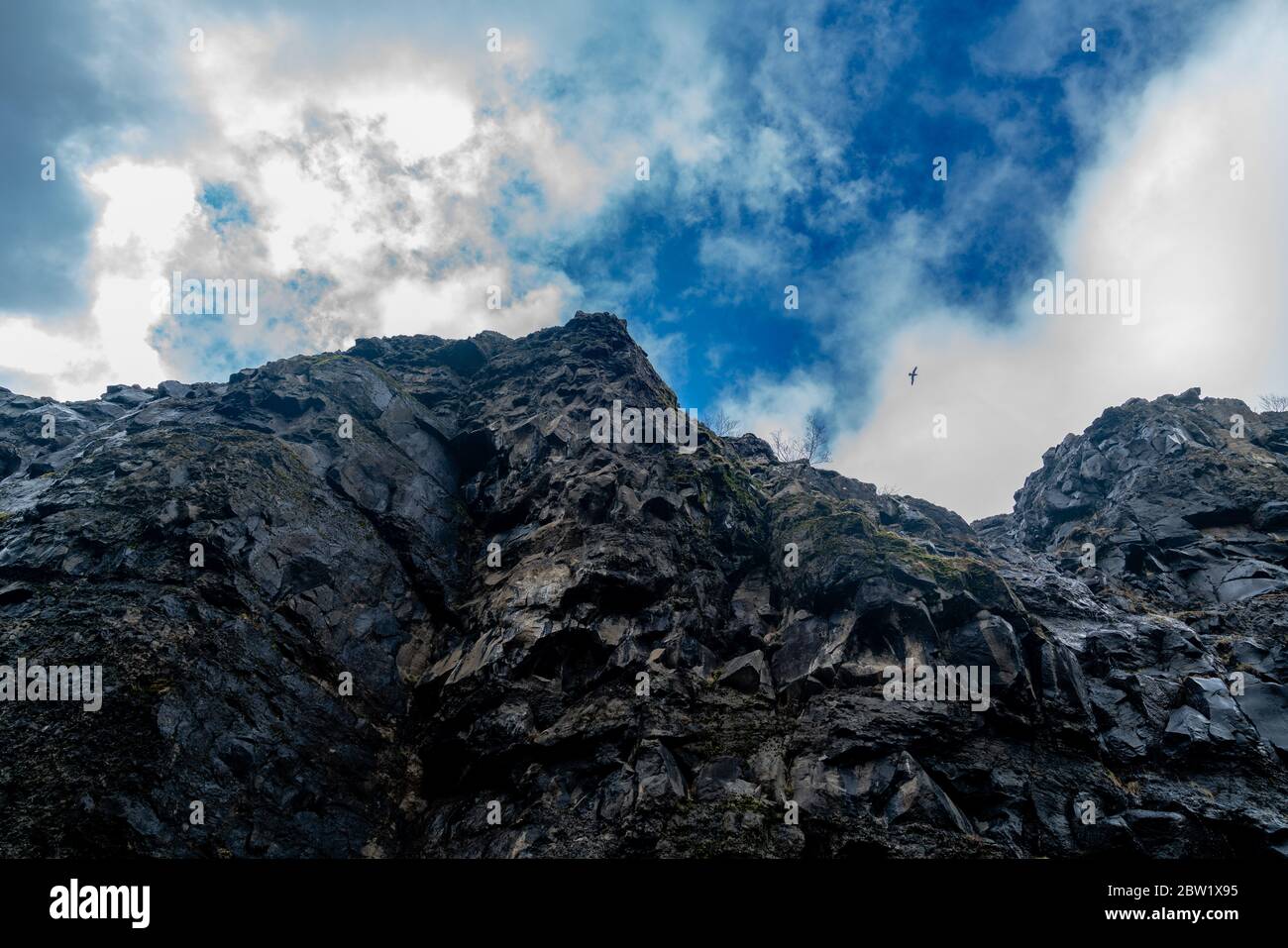 Ground view of a steep and rocky cliffside under a blue cloudy sky with a lonely bird flying Stock Photo