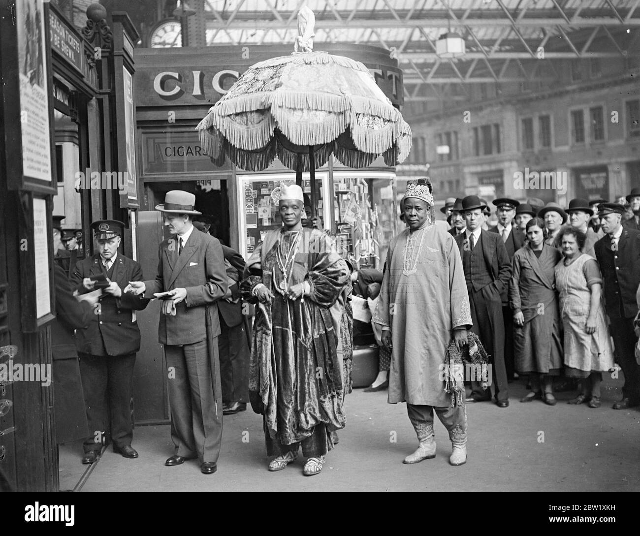 African ruler leaves thoughtfully to review under Royal umbrella. The Alake of Abeokuta, ruler of one of the largest tribes in Nigeria, left Waterloo station, London, to attend the fleet review at spit head. The Alake came to England to attend the Coronation, and is always sheltered by the Royal umbrella. Photo shows, the Alkake of Abeokuta under the Royal umbrella as he left Waterloo. 20 May 1937 Stock Photo