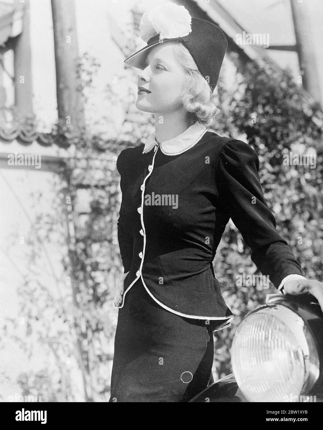 Mary Carlisle, the Hollywood film actress, wearing a new two-piece model of crepe [crêpe/crape] with a youthful tunic piped in white with a narrow silver edge. The buttons are tiny rhinestones, and a sash of the black crepe ties across the back. The hat is a smart black straw with a high crown surmounted by two feather pompoms. 7 May 1937 Stock Photo
