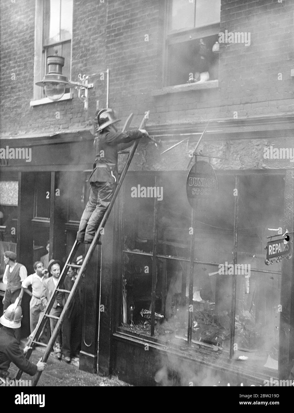A fireman on a ladder using an axe on the burning shop. Sea Scouts had to assist police to control the huge crowd which gathered in Lexington Street, Piccadilly, as firemen rescued a woman from a fire in cycle repair's shop. 27 August 1937 Stock Photo