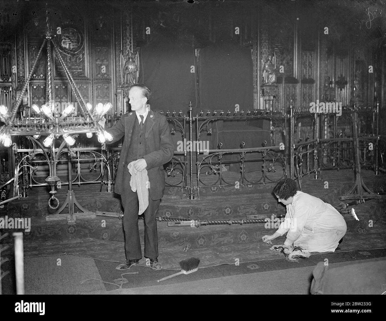 Mr A Atherton cleaning an electric light fitting as Mrs A Wheeler, who looks after all the carpets in the Houses of Parliament, stitching the carpet before the throne in which the King and Queen will sit in the House of Lords. The house is being made ready for the state opening of Parliament by the King and Queen on 26th of October when King George VI will perform the ceremony for the first time. 6 October 1937. Stock Photo