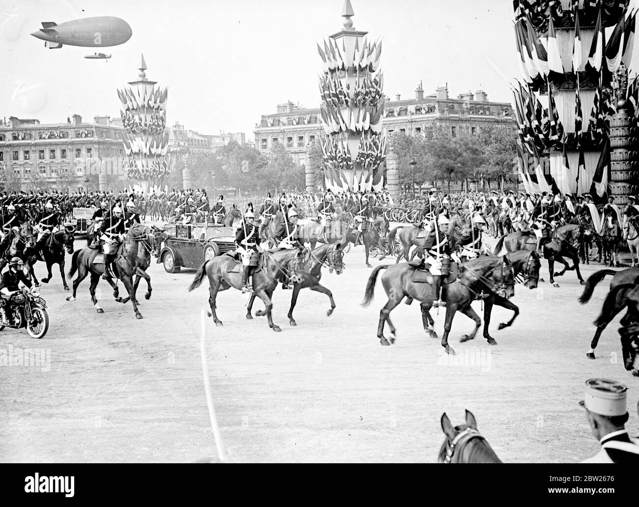 King and Queen receive tumultuous welcome in Paris. Welcomed by 1 million cheering people and the thunder of 101 gun salute, the King and Queen arrived at the Gare Bois de Boulogne on their State visit to Paris. Accompanied by President Lebrun of France and Mme Lebrun , they made the 4 mile drive through decorated streets lined with troops and crowds to the Qui d' Orsay Palace , where they are staying. Photo shows, the procession passing through the streets of Paris, escorted by cavalry and with a dirigible flying overhead.. 19 July 1938 Stock Photo