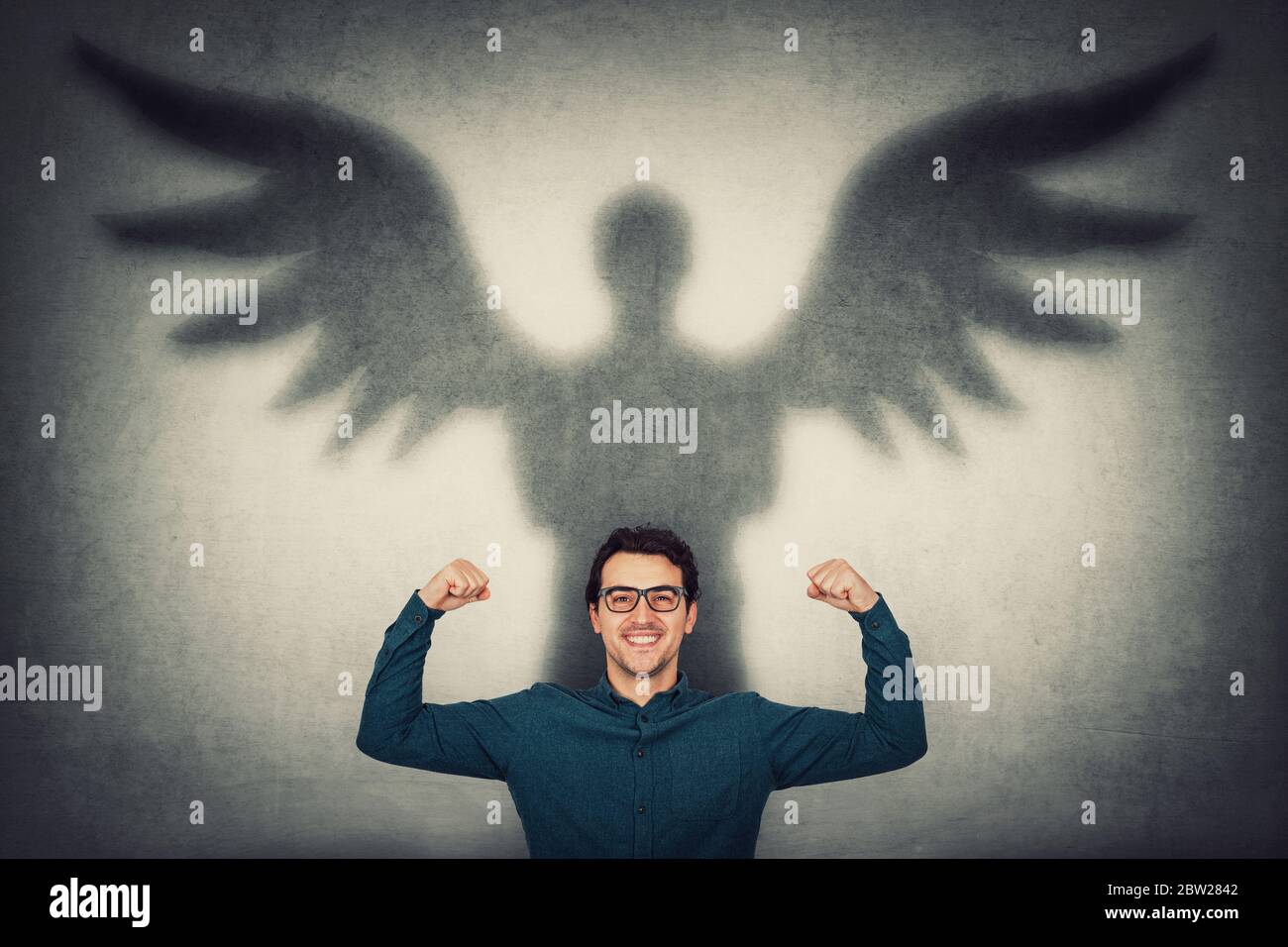 Confident businessman flexing muscles imagine superpower. Guy shows his strength, casting a superhero shadow with angel wings on a wall. Personal deve Stock Photo