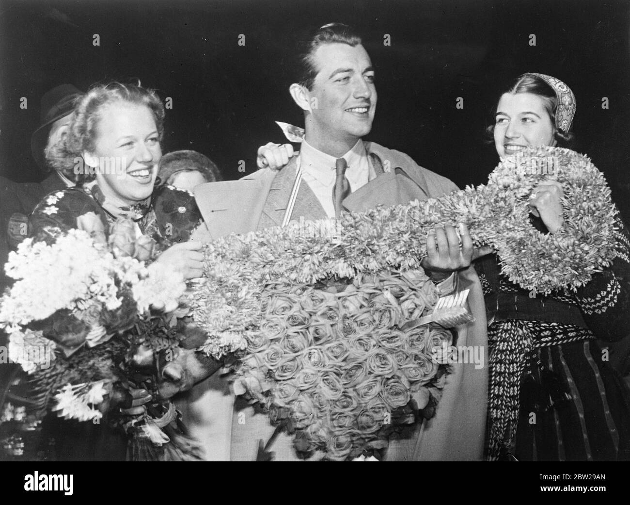 Feminine Sweden presents Robert Taylor, with the key to her heart!. A huge floral key and a heart of flowers were presented to Robert Taylor, the film actor, by two Swedish girls when he arrived at Bromma aerodrome, Stockholm. The girls were elected for the honour from hundreds of candidates. Photo shows, Robert Taylor, receiving the floral key and heart from the Swedish girls at Bromma Aerodrome, Stockholm. 26 November 1937 Stock Photo