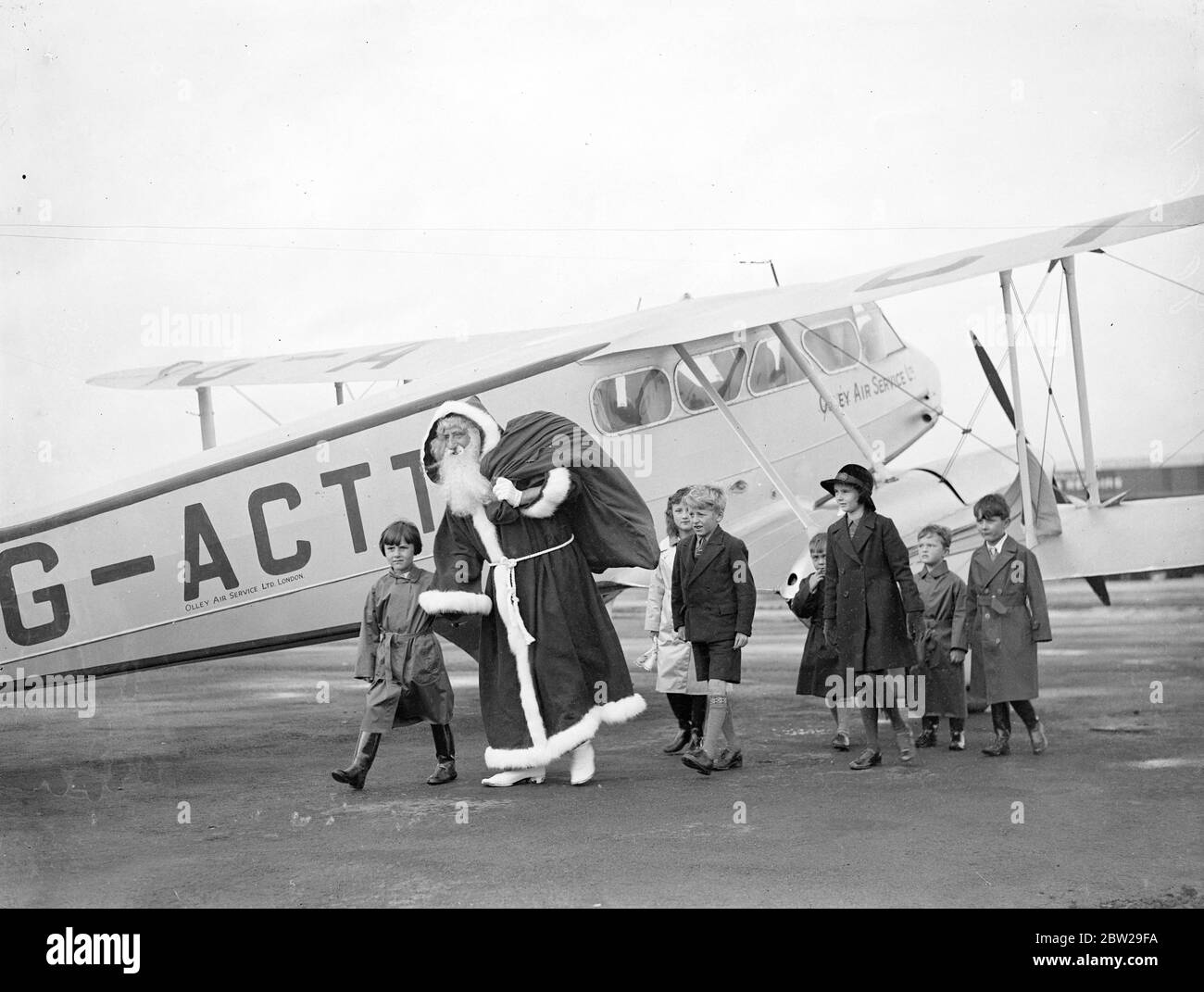 Father Christmas comes early for the rush. Arrives at Croydon by air. Discarding the usual sleigh and reindeer for more modern means of transport, Father Christmas, descended on London by air. He stepped from a plane at Croydon, to be greeted by cheering children who are already making their plans for Christmas, only eight weeks away. Photo shows, Father Christmas, carrying his sack, walking across the aerodrome, with children at Croydon. 25 October 1937 Stock Photo