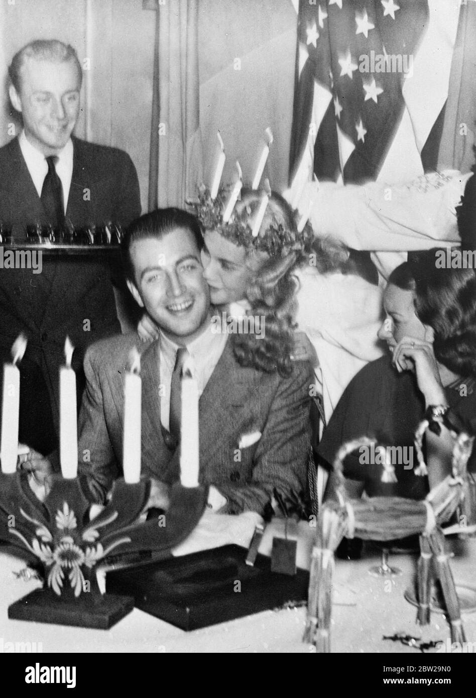 Robert Taylor receives kiss from a 'Lucia'. A lucky 'lucia' of Stockholm plants a kiss on the cheek of Robert Taylor, the film actor, after she had given him a glass of Swedish Bernt Brandy at a luncheon in his honour in Stockholm. The 'Lucia' is Sickan Carlsson, Swedish film actress. She is wearing the traditional Crown of lighting candles with which St Lucia's Day (13 December) is welcomed in Swedish homes. In accordance with the custom, a feminine member of each family is chosen to be the 'Lucia'. The lighted candles symbolise the returning sunlight. 28 November 1937 Stock Photo