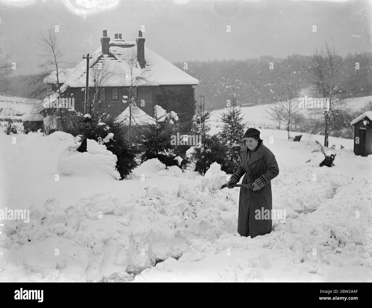 Digging her gardens out of the snow in Hampshire. The worst December snowstorms for many years had brought chaos to southern England, where many villages had been cut off by snow drifts. Cars have been abandoned by their drivers and left on the roads to be almost buried by the snow. Photo shows, a 'Christmas card' scene near Ringwood, Hampshire, where a girl is clear and deep snow from the garden of a picturesque cottage. 9 December 1937 Stock Photo