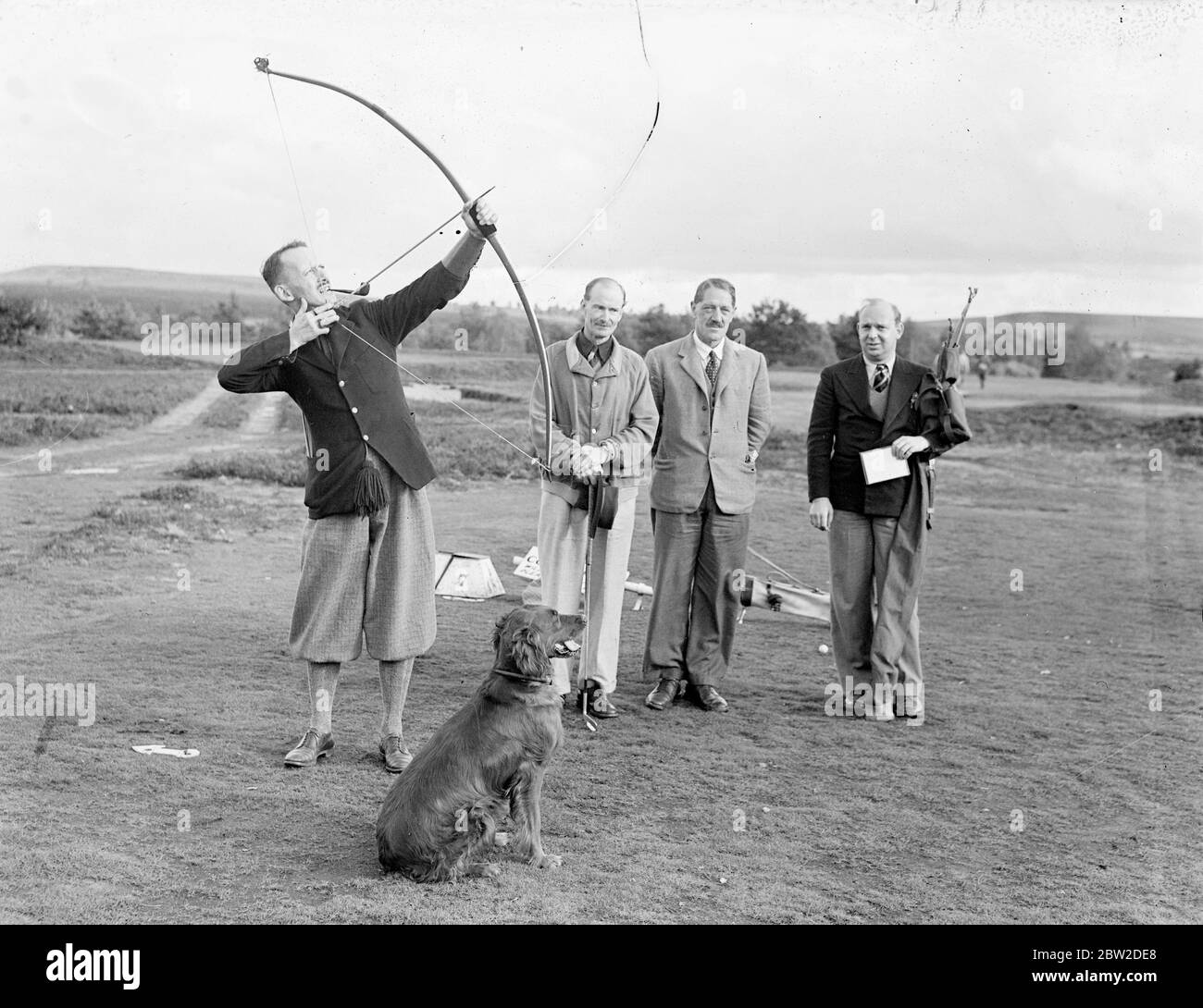 Playing with bow and arrow, Major J G Hayter, acknowledged expert at the annual sport of bow and arrow golf, met Colonel E St George Kirke, the captain of the club, in a match at the Hankley Common Golf Club, Surrey. He conceded a stroke a gale. Major Hayter shoots his arrow into a box instead of a hole. His average for most of the Surrey courses is in the seventies. Photo shows: Major J G Hayter shooting from the first tee while his pet Irish Settler dog Becky watches the flight of the arrow.. 18 October 1938 Stock Photo