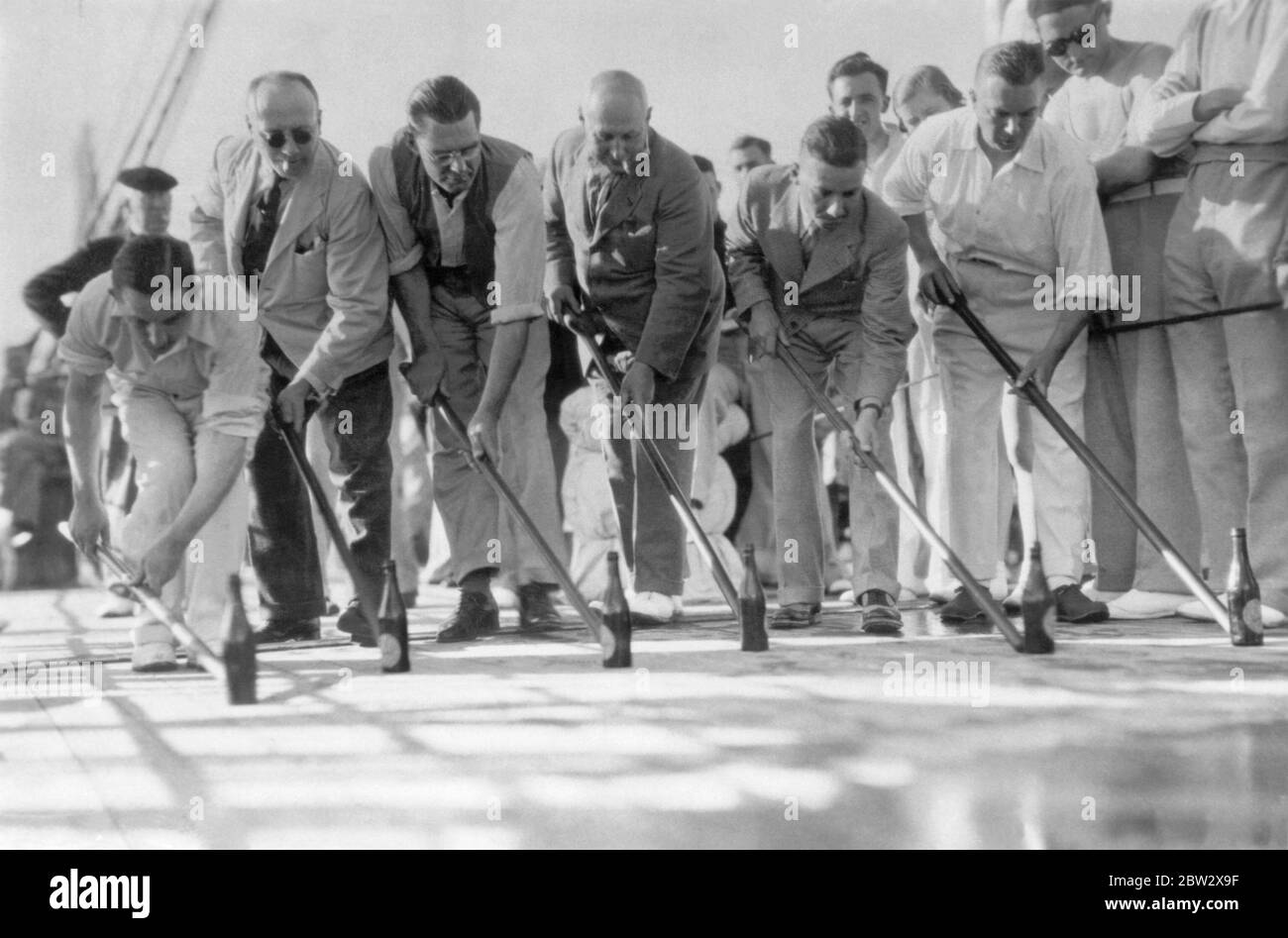 Deck games on board an ocean liner in the 1930s. Here is a variation on the game of 'Shuffleboard' with a race involving empty beer bottles being pushed along by six male passengers using long sticks. Stock Photo