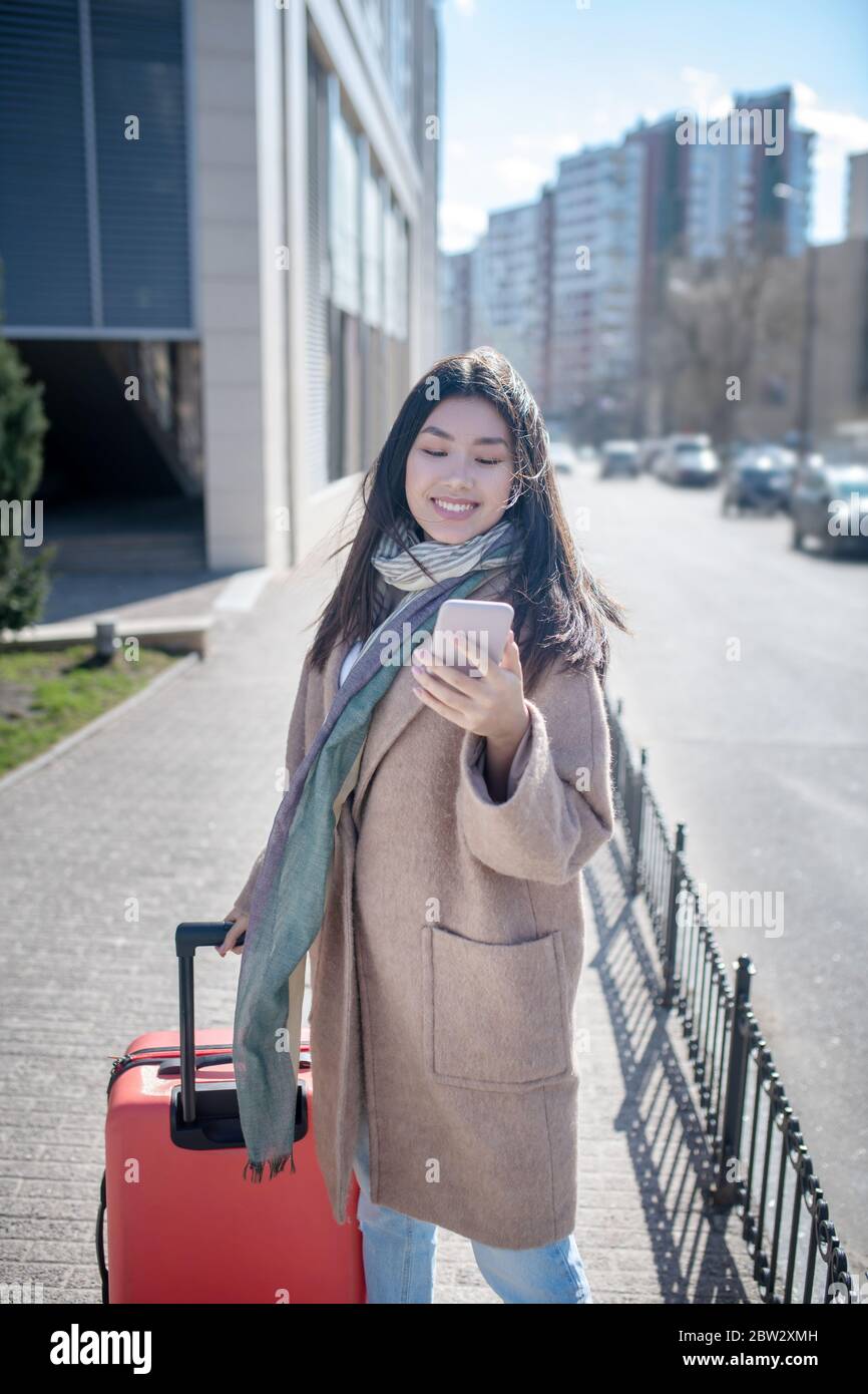 Brunette female in beige coat walking along the street with red suitcase, checking her mobile Stock Photo