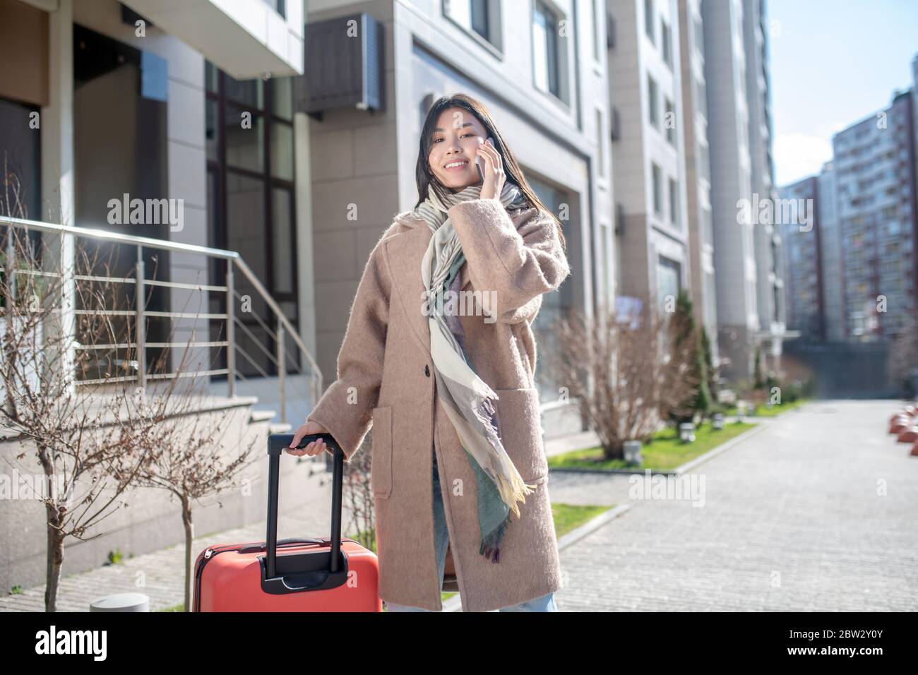 Brunette female in beige coat walking along the street with red suitcase, talking on her mobile Stock Photo