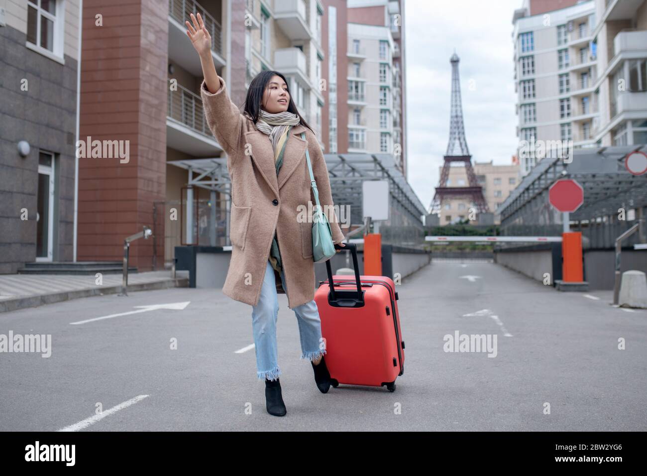 Brunette female in beige coat walking with red suitcase, trying to catch taxi Stock Photo