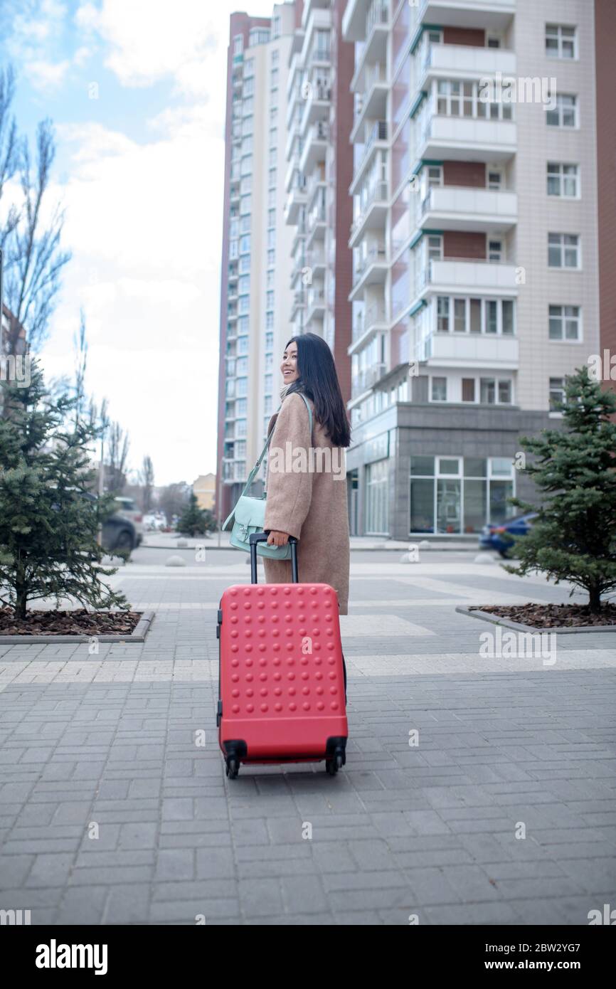 Brunette female in beige coat walking with red suitcase along the street Stock Photo