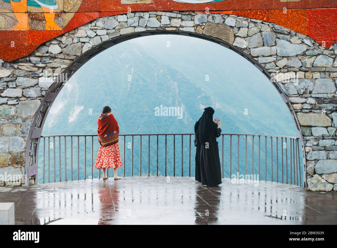 Two tourists stand under a stone arch on the Russia–Georgia Friendship Monument in the Caucasus Mountains, Georgia Stock Photo