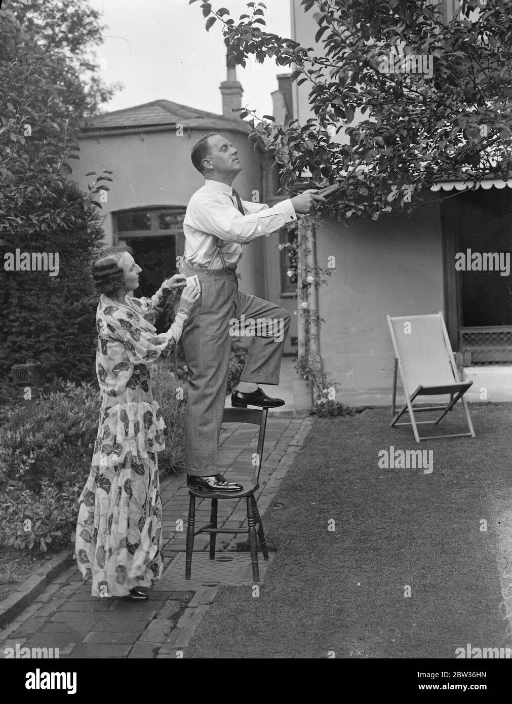 Gillie Potter , the famous radio comedian , being supervised by his wife whilst doing a spot of gardening . Stock Photo