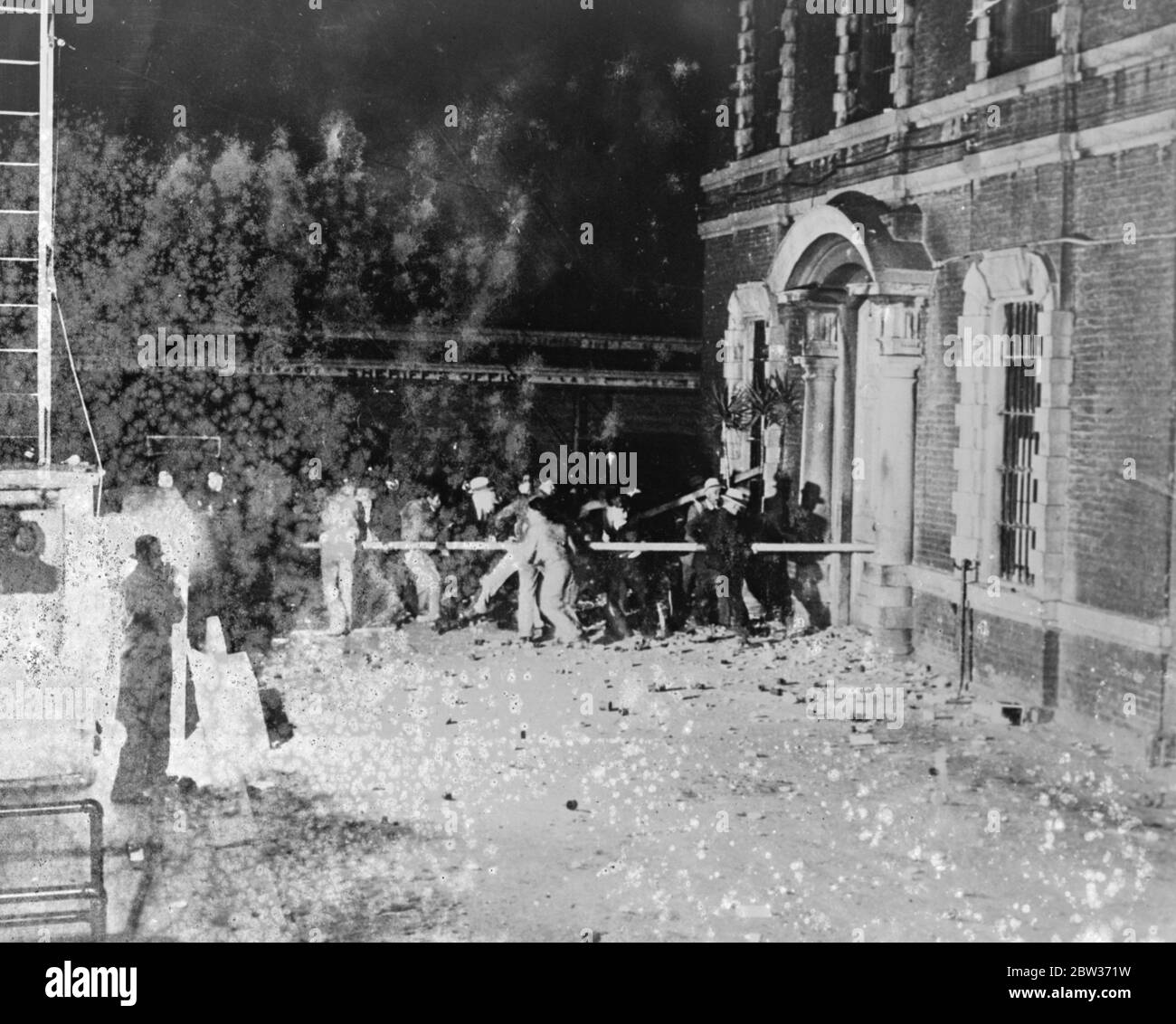 A mob using improvised rams , battered a way into the gaol at San Jose , California , to remove John M Holmes and Thomas H Thurmond , who had confessed to the kidnapping of Mr Brooke Hart , who was found murdered . Bricks and stones hurled by the furious crowd flew constantly at the building and tear gas was used in reply . The two men were eventually dragged from the prison and hanged from trees . Phohto sows ; Part of the mob battering down the doors of the gaol at San Jose , California . A cloud of tear gas is hovering over the building in centre of picture . 6 December 1933 Stock Photo