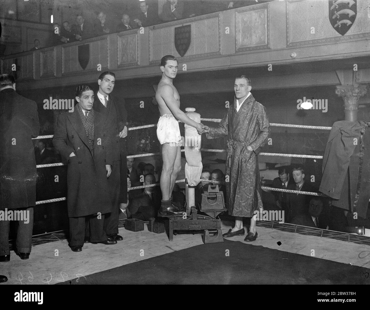 Watson and Crowley weigh - in for big fight . Seaman Watson , the Featherweight Champion of Britain , and Dave Crowley of Clerkenwell , weighed - in at the Stadium Club , Holborn , for big fight at Olympia . Photo shows ; Crowley ( on scales ) shaking hands with Seaman Watson , at the weigh in . 19 December 1933 Stock Photo