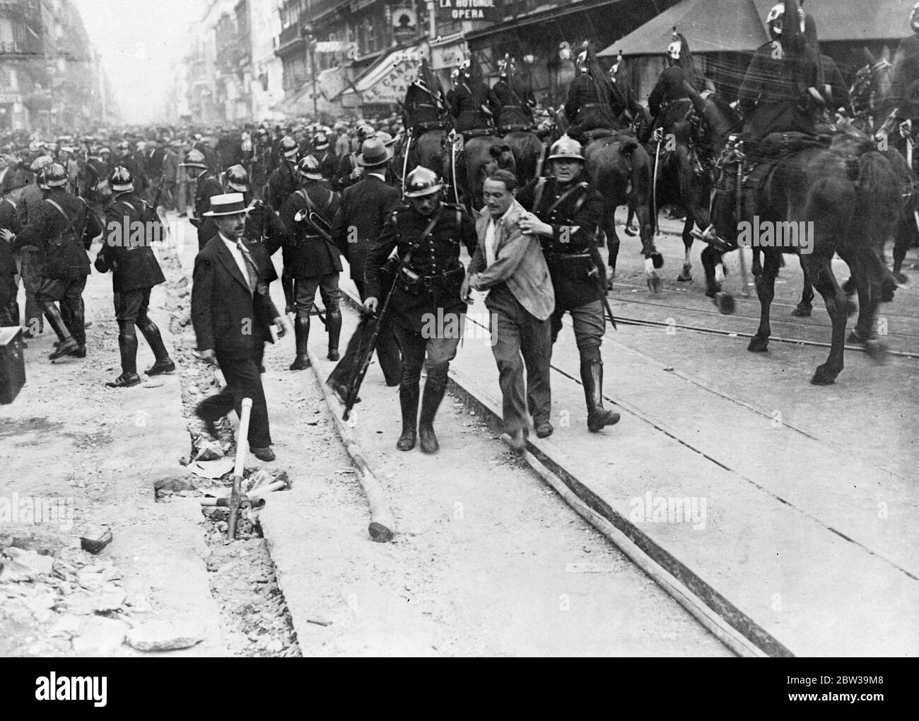 More than a thousand arrests in Paris anti wage cut riots - police in fierce battles with demonstrators . A demonstrator being led away by Mobile Guards in the Rue Lafayette , as mounted troops charge the crowd in background . 20 July 1935 Stock Photo
