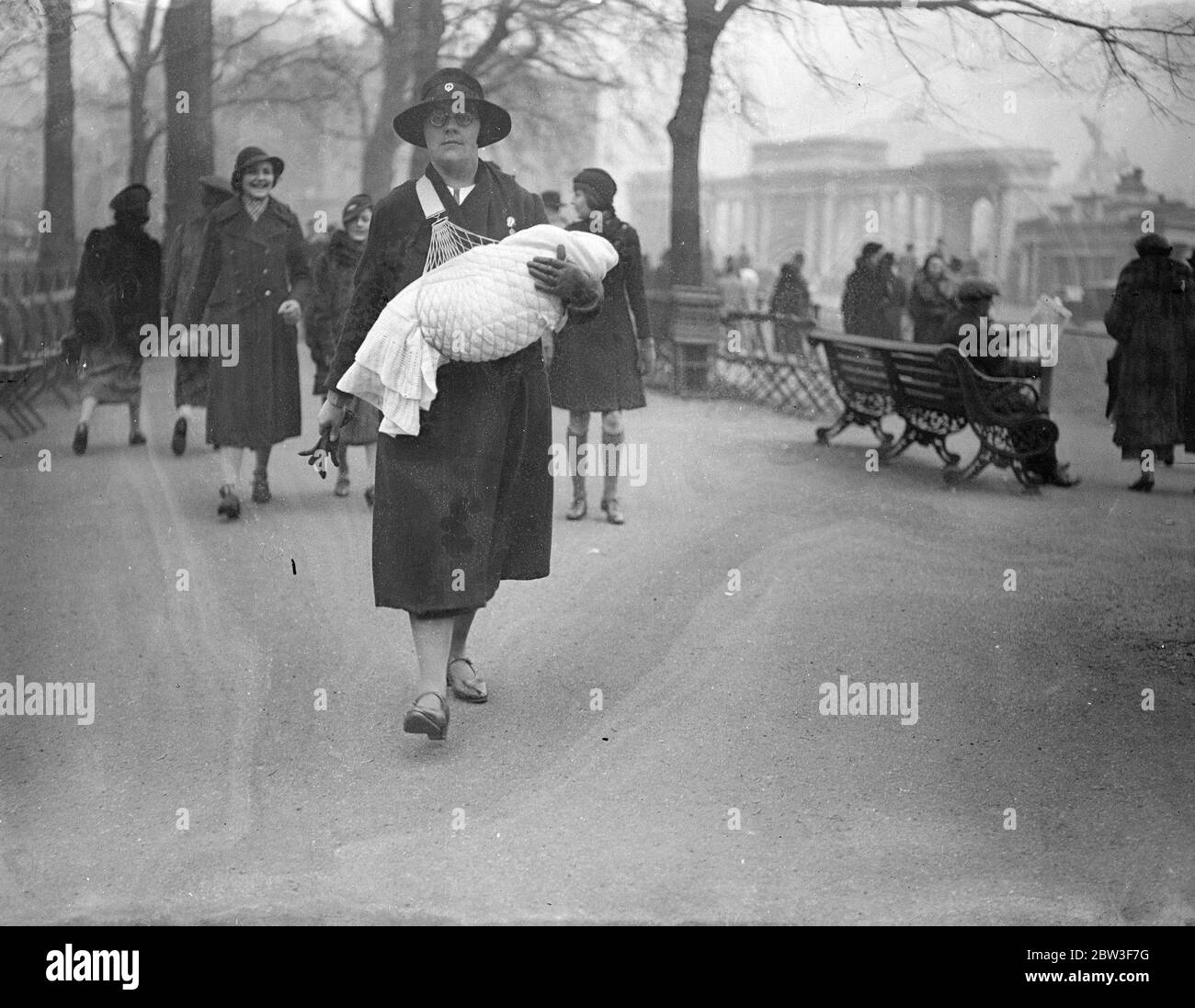 Carrying the baby in comfort . A London nursemaid has an ingenious device to relieve arm strain when she takes her charge for his morning walk in the Park . the baby is carried in the arms in the ordinary manner , but is suspended in a strong ' hammock ' , which is in turn attached to a broad strap running over the shoulder and under the arm . The weight is evenly distributed over the nurse ' s arms and shoulders and there is no sagging on the part of the baby , who rests quite comfortably and easily in the hammock . Photo shows , taking the baby for his morning walk , using the new device , i Stock Photo