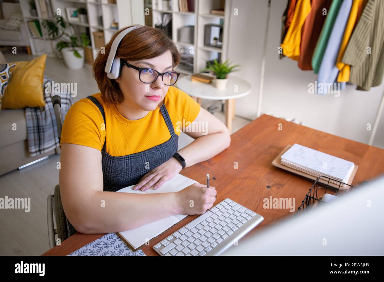 Focused young woman in wireless headphones sitting in wheelchair and making notes about lecture while studying at home Stock Photo