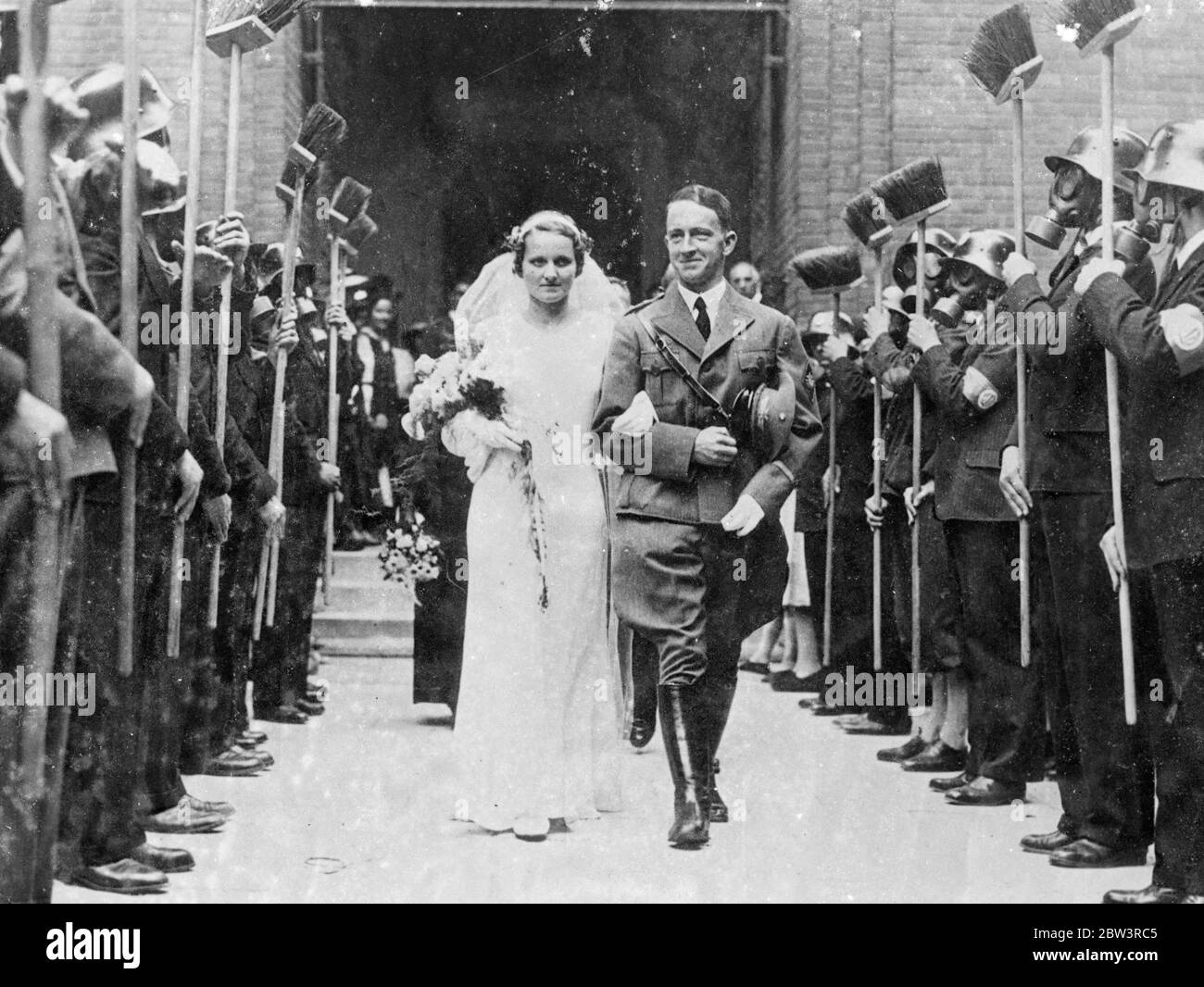 First  Air Defence  Wedding In Vienna , Gas - Masked Guard Of Honour The guard of honour wearing gas - masks and steel - halmets and holding lime brooms attended the first  air - defence  wedding in Vienna . Air Defence Leader Hugo Noll was married to Fraulein Helga Schenk . The strange guard of honour  presented brooms  as the bridal couple left church . Photo shows : The bride and groom walking through the guard of honour of gas - masked anti air - raid men . 4 Jun 1936 Stock Photo