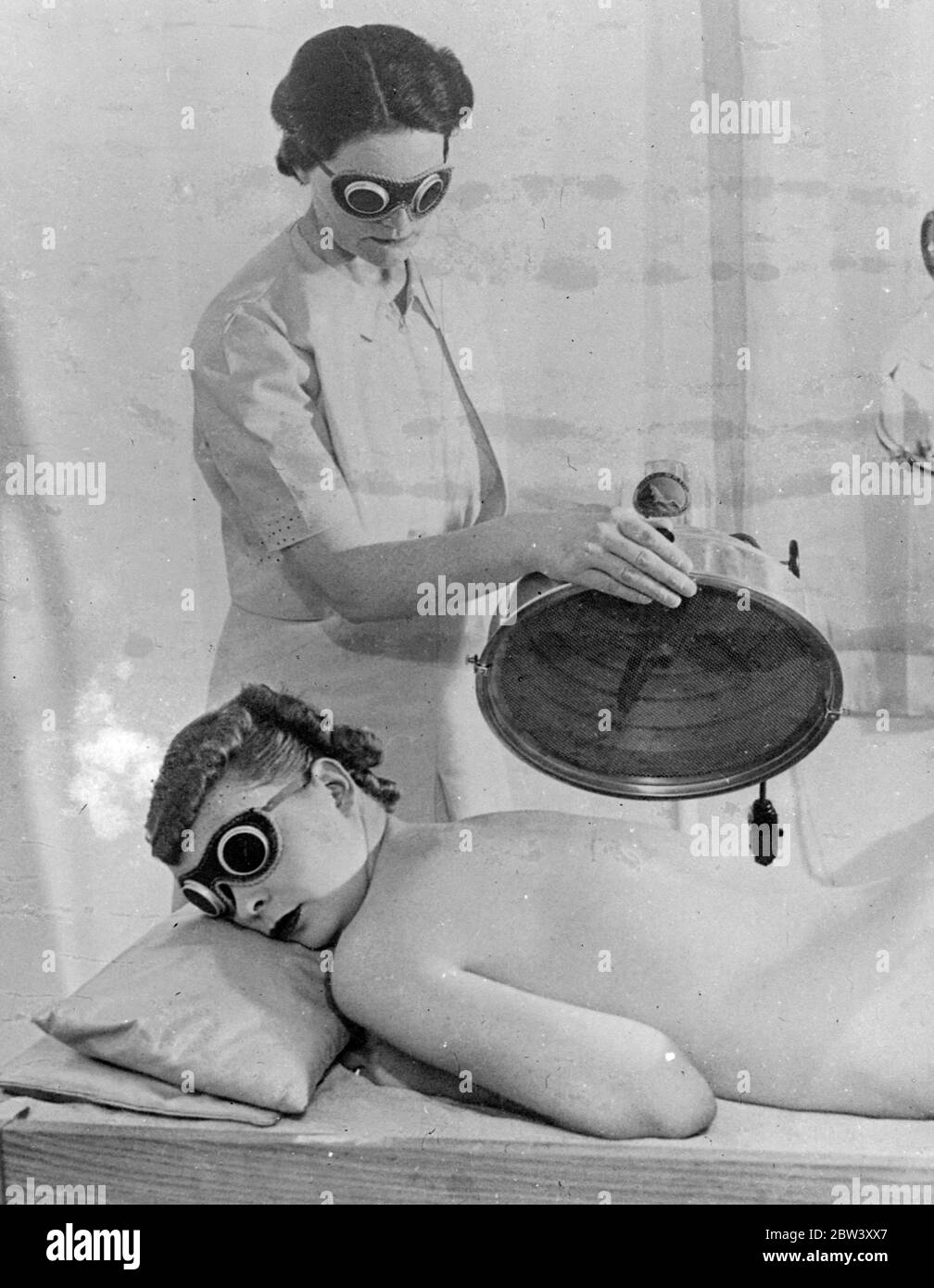 Beauty by ' Mass Production ' methods . Beauty treatment has been organised on the lines of ' Big Business ' in America and 'mass production ' methods introduced to the salons . Though the client is not actually placed on a moving belt , she moves from one department to another for each special treatment , as this series of pictures , made in a Fifth Avenue , New York , salon , shows . Photo shows , Ultra violet ray treatment , one of the most healthful aids to beauty , for a client at the New York salons . The whole room is bathed in the light and both the girl , who is reclining on real sand Stock Photo