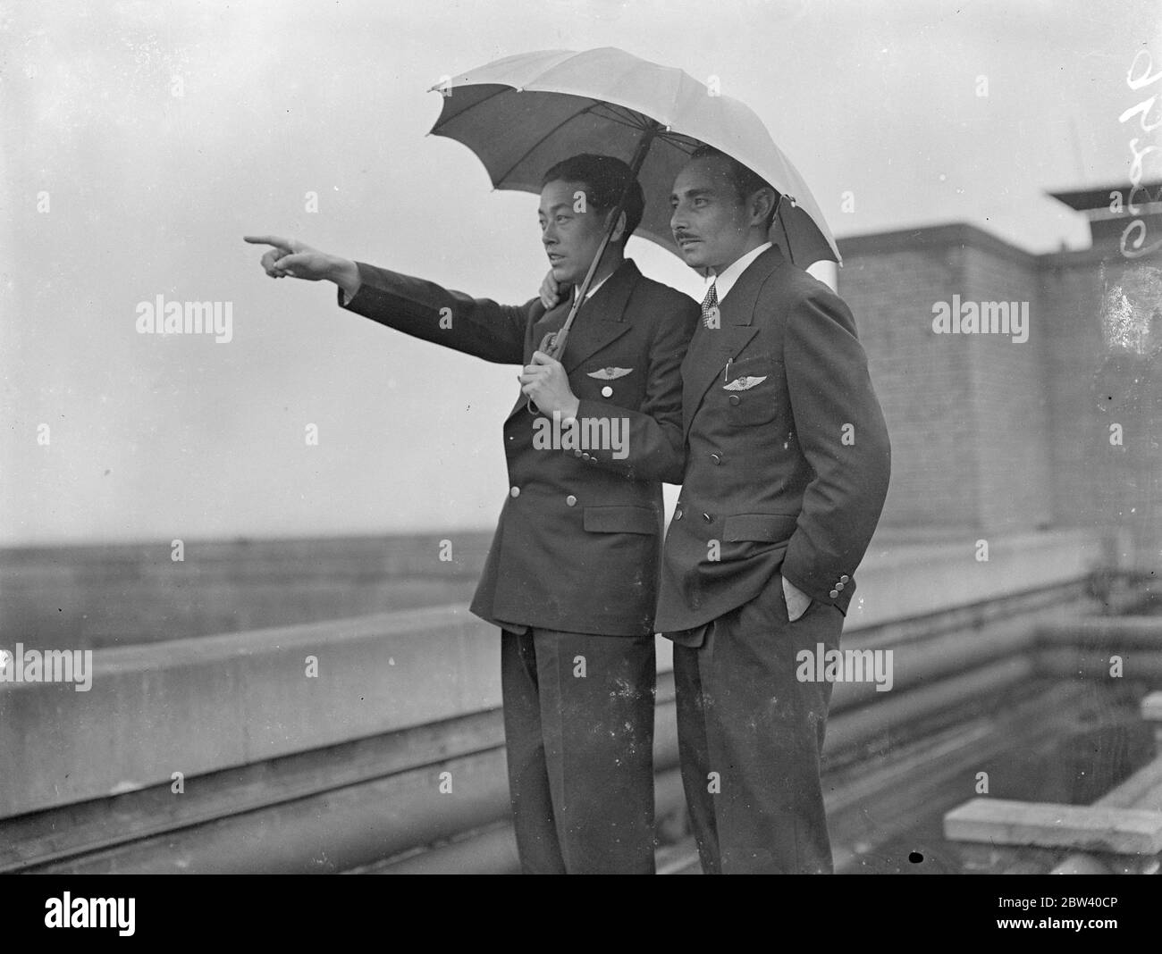 So this is London! Japanese airmen take a closer look in the rain. After smashing all records by flying the 10,000 miles from Tokyo to London in well under 100 hours, the Japanese airmen Masaaki Iinuma and Kenji Tsukakoshi, landed Croydon in their plane Divine Wind [Mitsubishi Ki-15 Karigane aircraft, name: Kamikaze, registration J-BAAI]. The flyers have brought Coronation greetings from Japan. Photo shows: sheltering under an umbrella the Japanese airmen Masaaki Iinuma (left) the 27-year-old pilot, and his radio operator Kenji Tsukakoshi get closer look at London from the roof of their hotel. Stock Photo