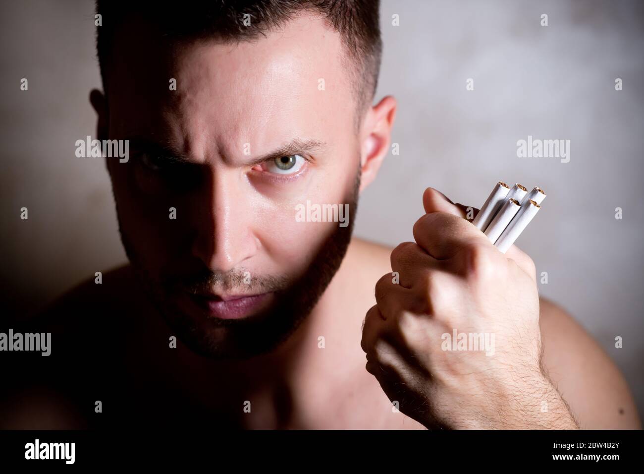 unshaven man stares sternly at the camera, clutching cigarettes in his fist. close up, side lighting, contrast light Stock Photo