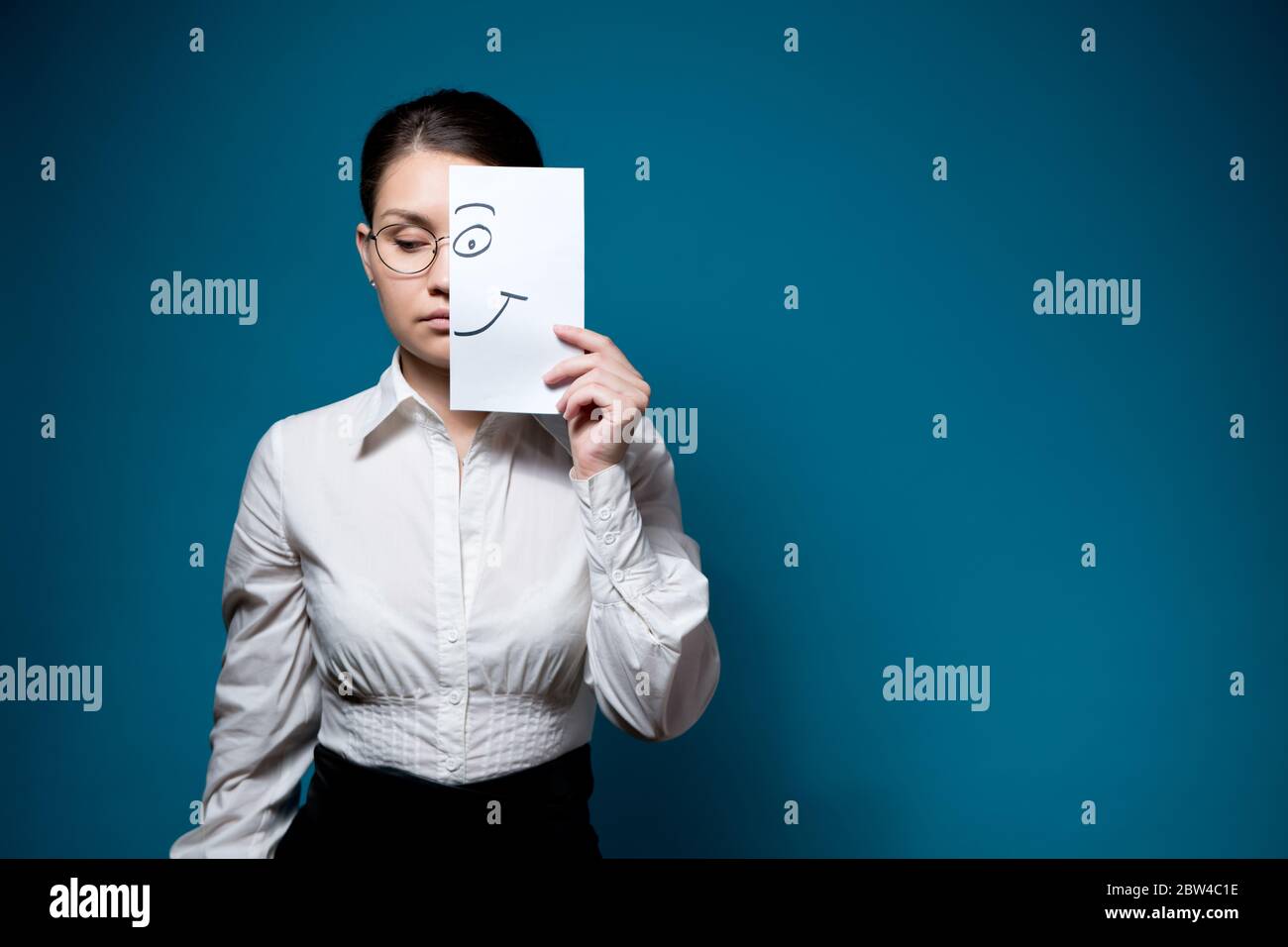 unemotional woman with glasses and a white shirt hides her eyes and covers half her face with a piece o paper with a painted smile Stock Photo