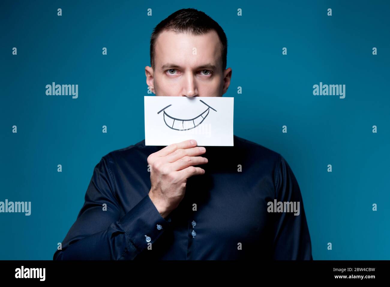 unemotional man in a blue shirt holds a tablet with a painted smile near his mouth Stock Photo