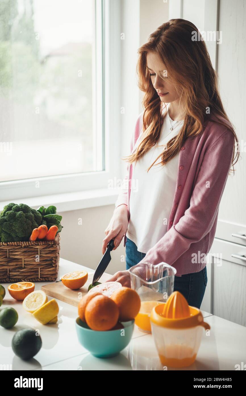 Caucasian fitness mother with red hair and freckles cutting fruits and making natural fresh juice in the kitchen from oranges and lemon Stock Photo