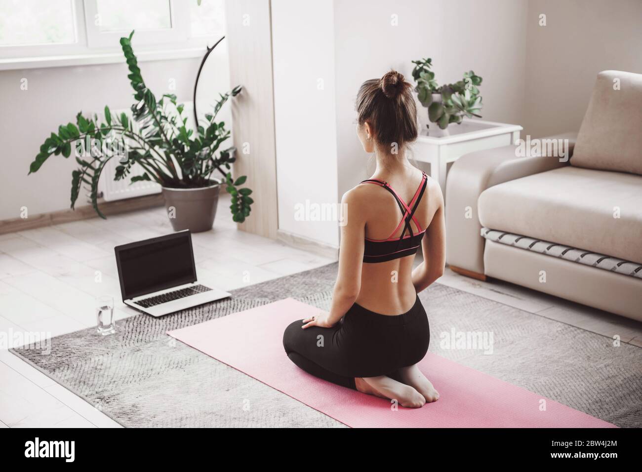 Caucasian woman sitting in front of the laptop and practice yoga at home dressed in sportswear on a pink sport carpet Stock Photo