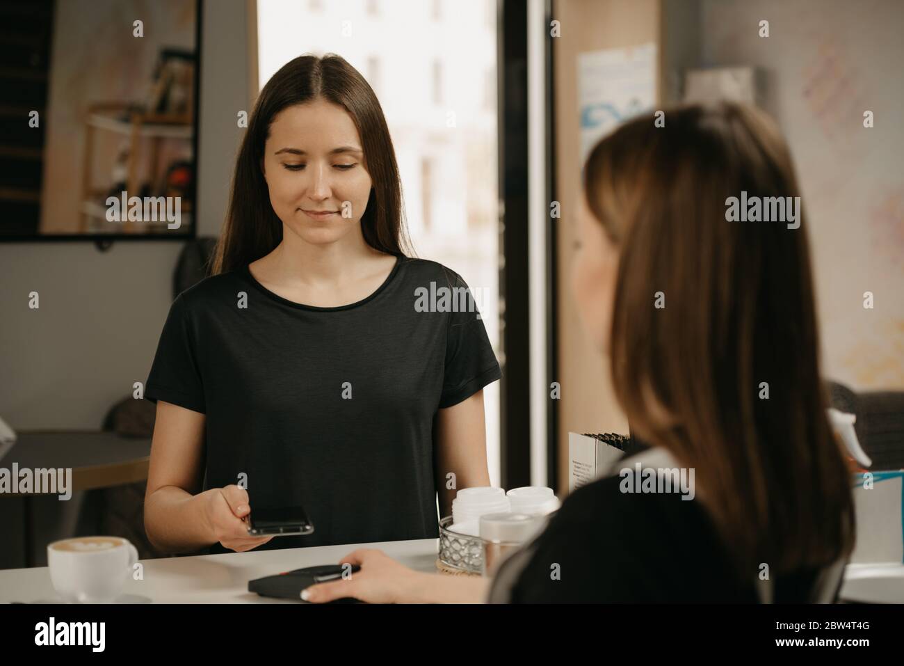 A girl with long hair paying for her coffee with a smartphone by contactless NFC technology in a cafe. A female barista holds out to a client a termin Stock Photo