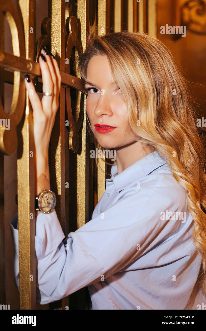 Portrait of a beautiful girl with long curly blonde hair, wearing a light top. She is is next to a fence, holding it with a hand. She's looking at us Stock Photo