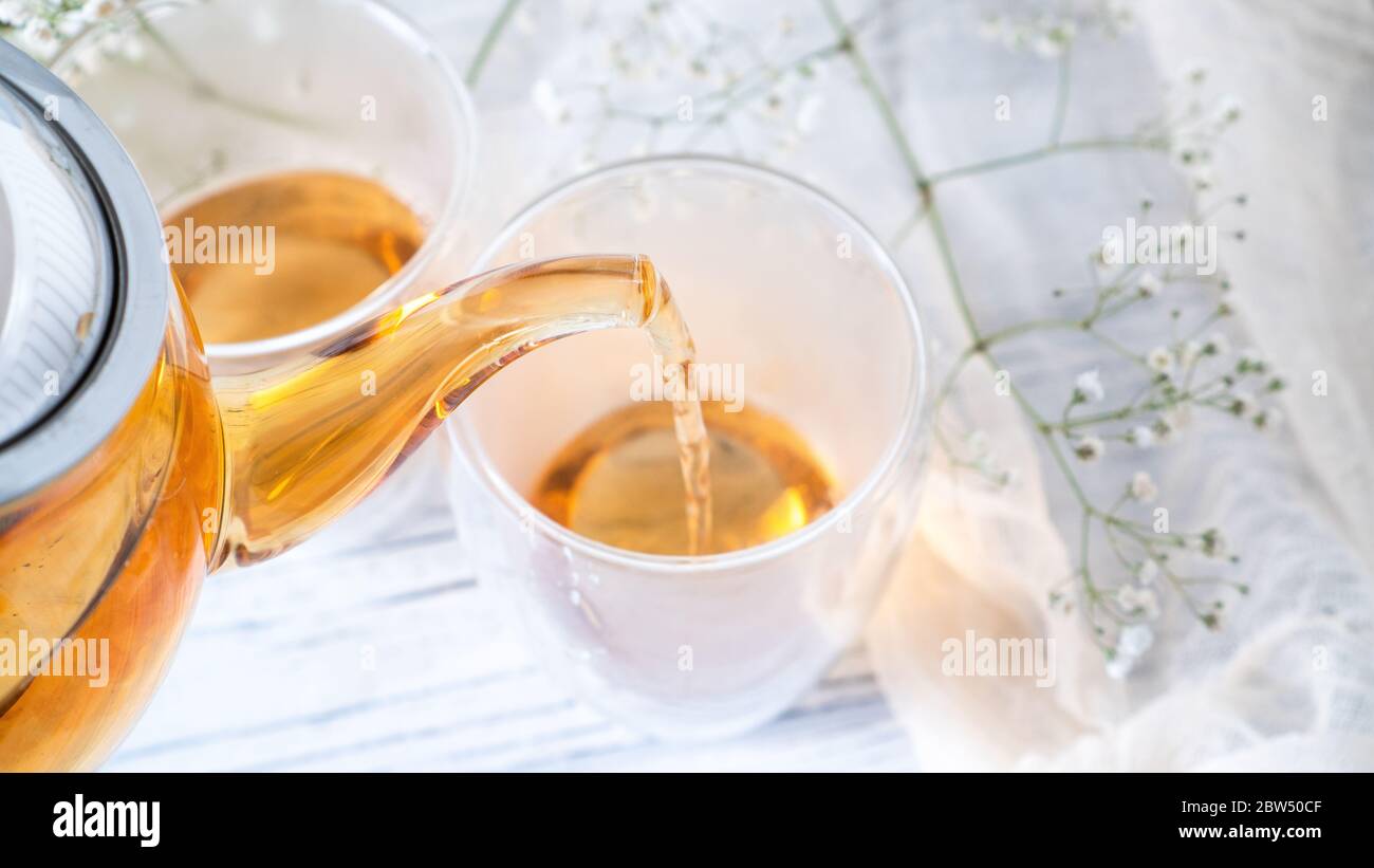 From transparent glass teapot pour golden tea in glass mug. Glass teapot pouring black tea into cup. White wooden table Stock Photo