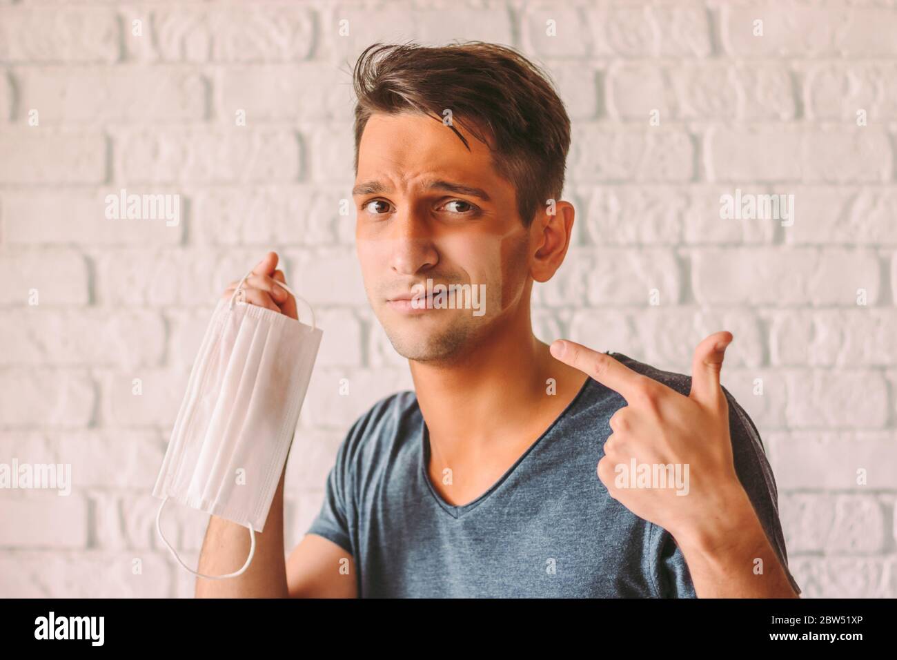 Portrait confused man with funny sun tan lines hold protective mask in hand. Young hipster man with red sunburn skin after wearing medical face mask. Stock Photo