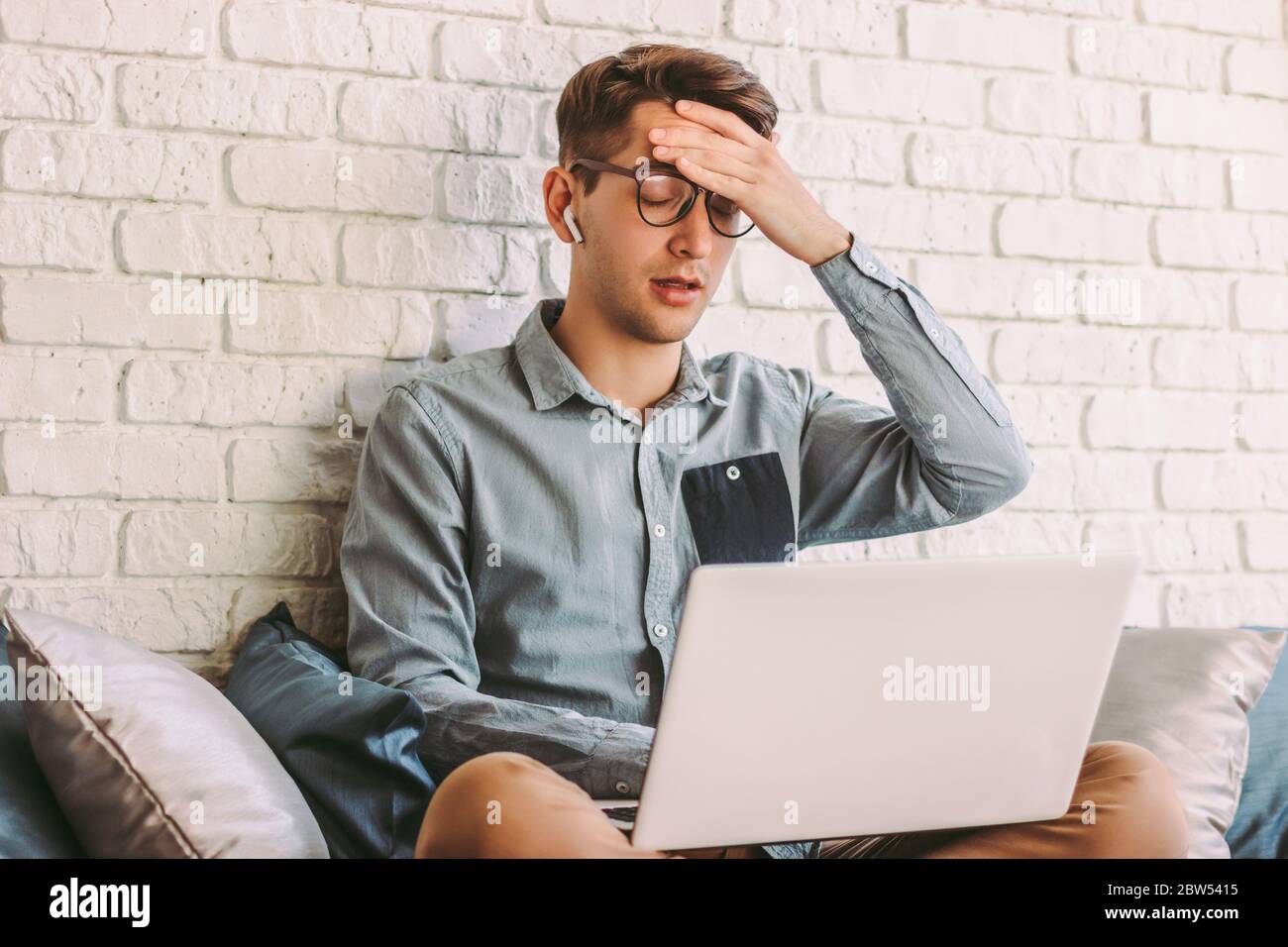 Young stressed hipster man in glasses feel tired, exhausted while use laptop on couch. Nervous frustrated freelance businessman look unhappy or depres Stock Photo