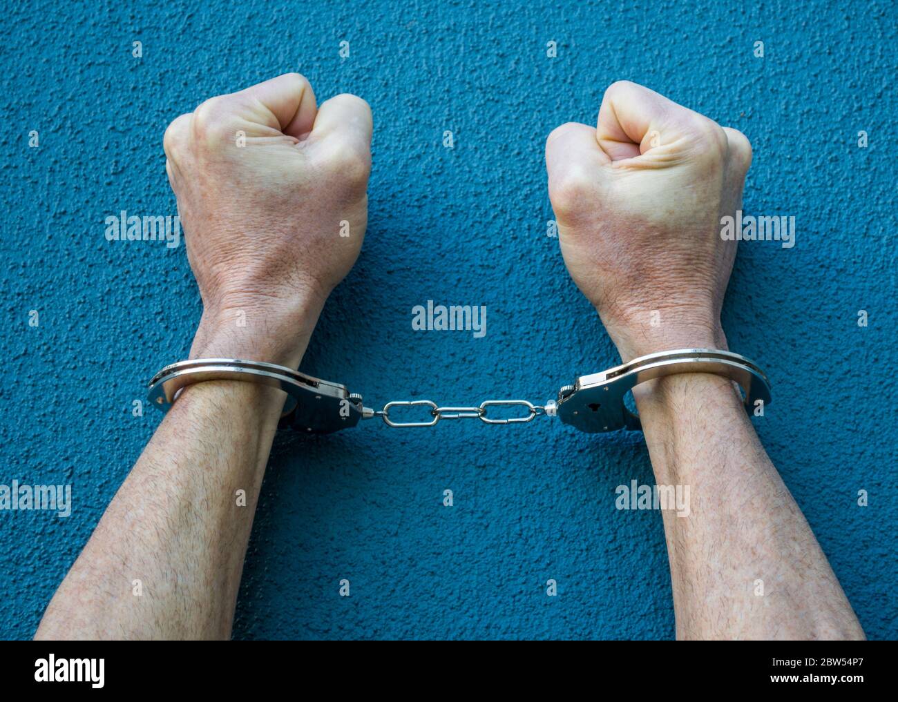 arms and fists of a Caucasian man in handcuffs against a blue background with copy space Stock Photo