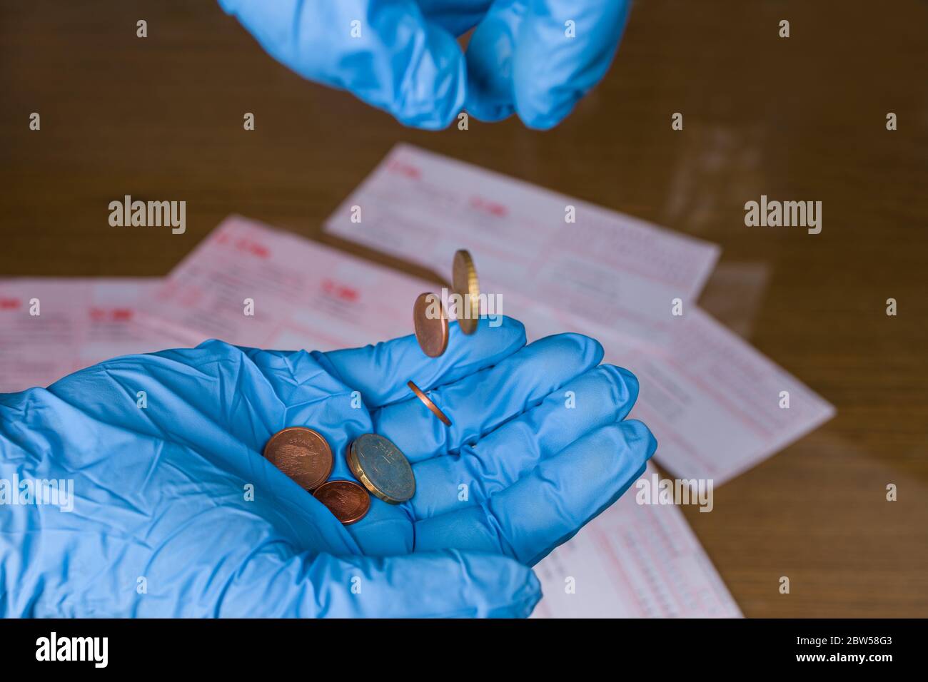 Euro coins falling to palm in blue protective glove, money orders on wood desk. Economic and financial loss. Poverty hardship in coronavirus pandemic. Stock Photo