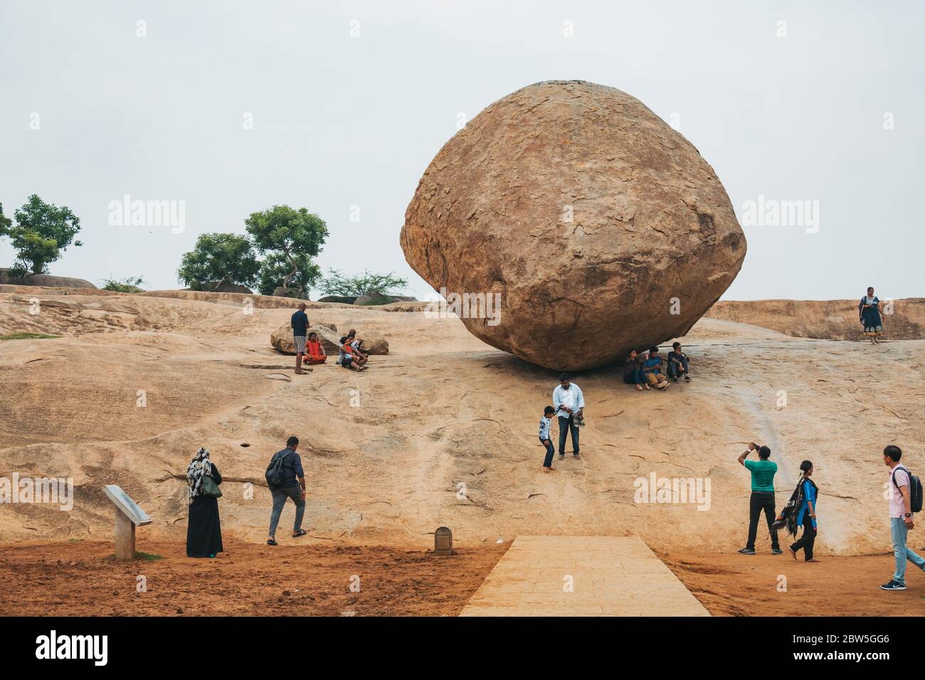 Tourists sit under Krishna's Butter Ball, a huge granite boulder perched on a downhill slope in Mahabalipuram, Tamil Nadu, India Stock Photo