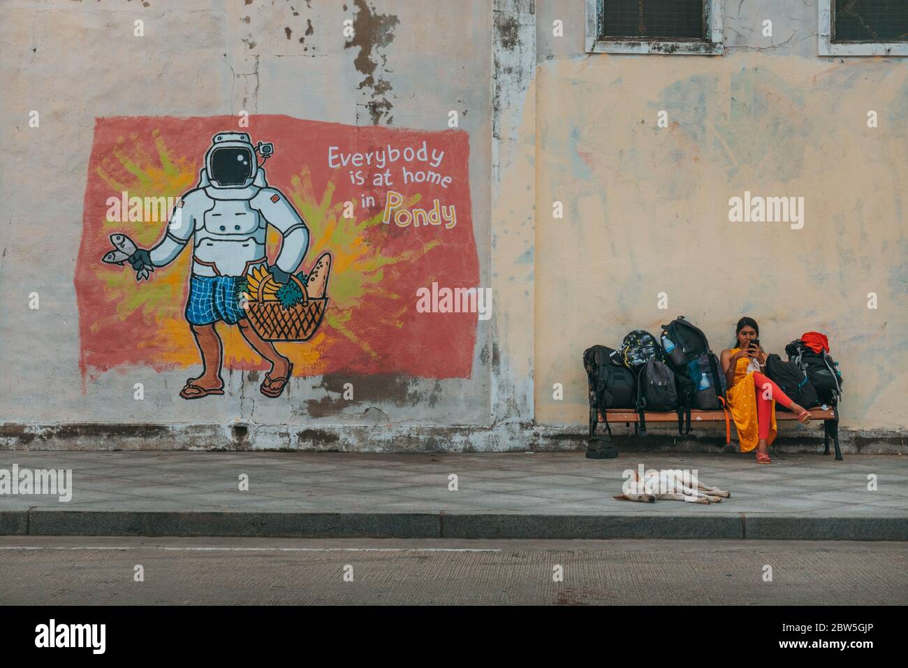 A woman waits with luggage on a bench next to street art with the text 'Everybody is at home in Pondy' in Pondicherry, India Stock Photo
