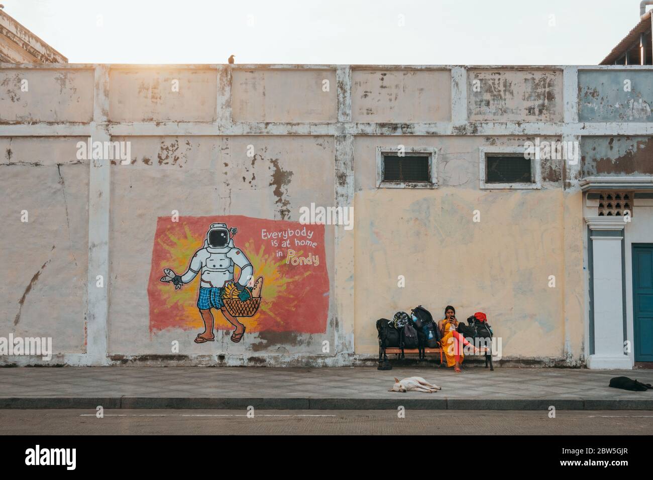 A woman waits with luggage on a bench next to street art with the text 'Everybody is at home in Pondy' in Pondicherry, India Stock Photo