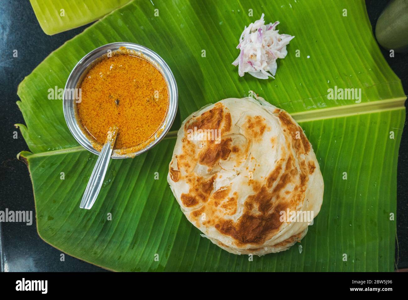 Parotta (flaky flatbread) served on a leaf with onion and a bowl of sambar in Munnar, Kerala, India Stock Photo