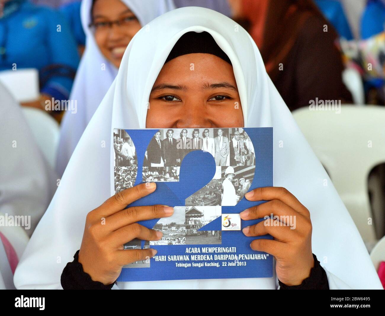 Malay Muslim girl with smiling eyes wears an Islamic school uniform and covers her mouth with a patriotic brochure. Stock Photo